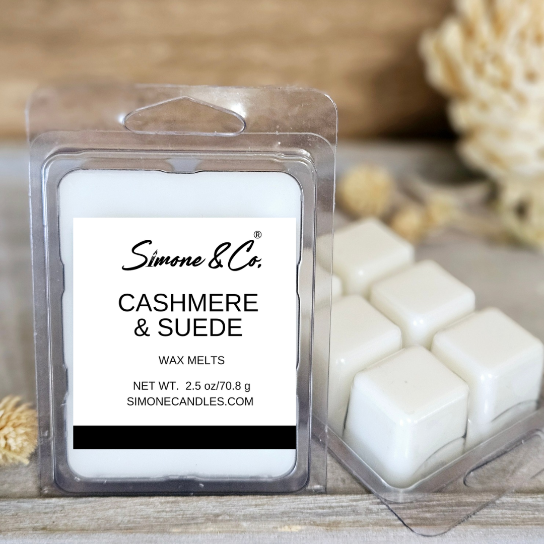 A package of Simone & Co. Cashmere & Suede wax melts in a clear clamshell tray, shown next to several white wax melts in cube shapes on a rustic wooden surface with soft beige dried floral props in the background.