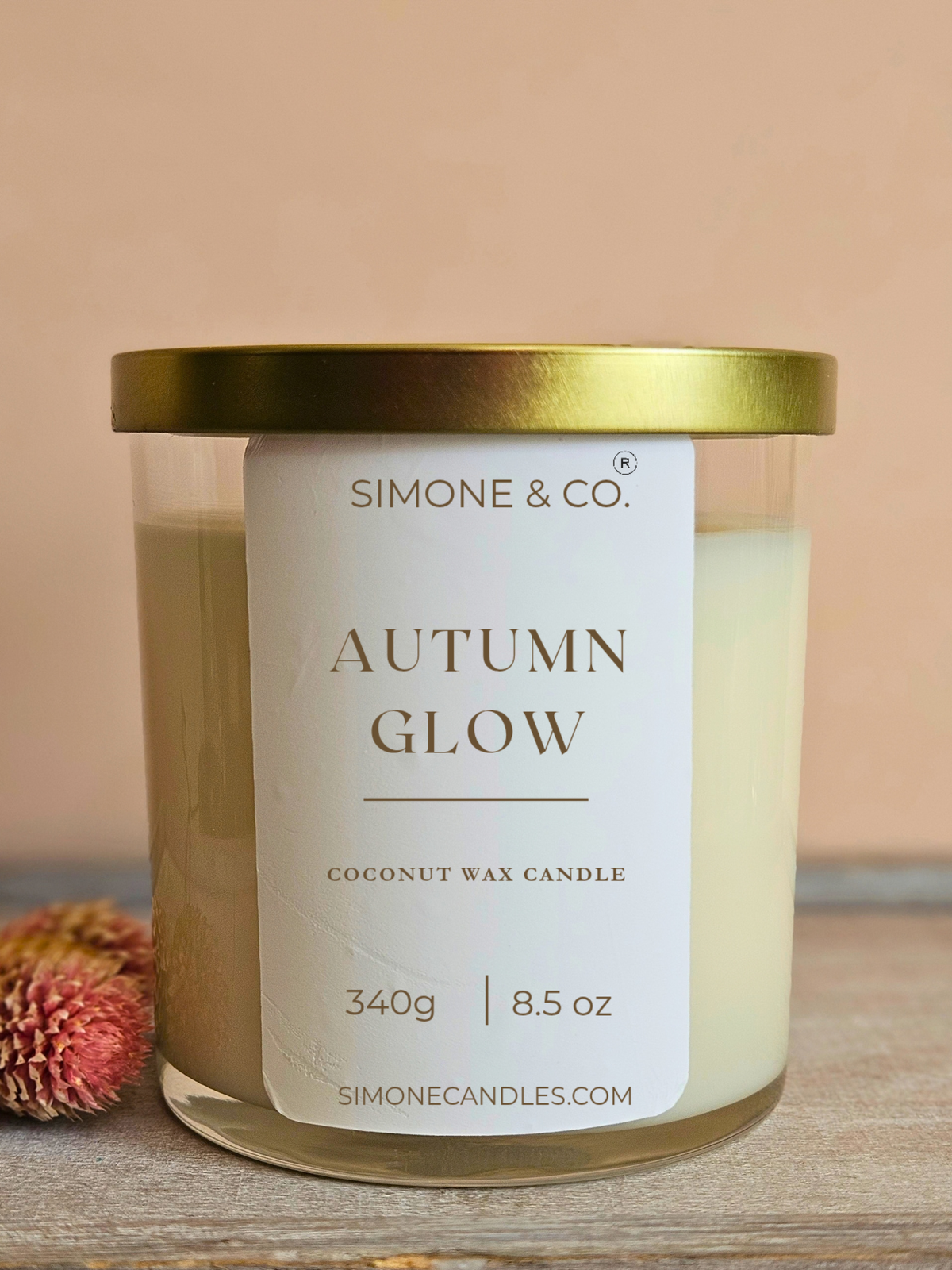 A candle in a clear glass jar with a white label reading 'SIMONE & CO. AUTUMN GLOW' and a metallic gold lid, set on a wooden surface with a soft beige background and a small dried flower prop.