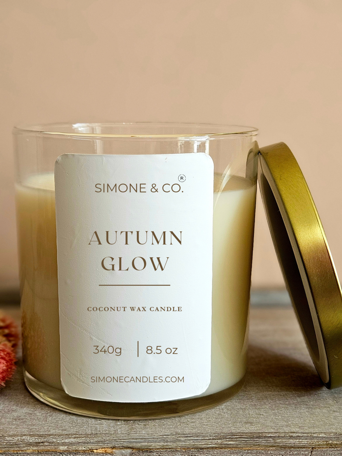 A candle in a clear glass jar with cream-colored wax and a white label reading 'Simone & Co. Autumn Glow Coconut Wax Candle.