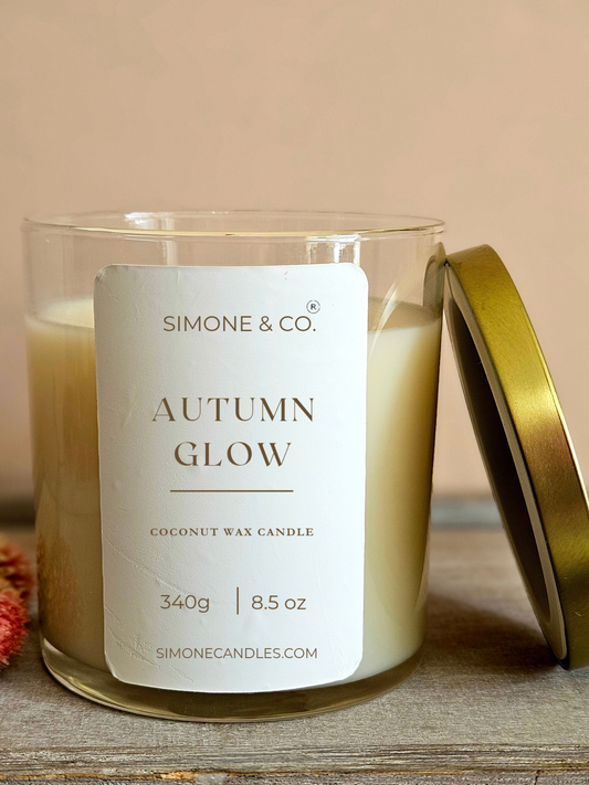 A candle in a clear glass jar with cream-colored wax and a white label reading 'Simone & Co. Autumn Glow Coconut Wax Candle.