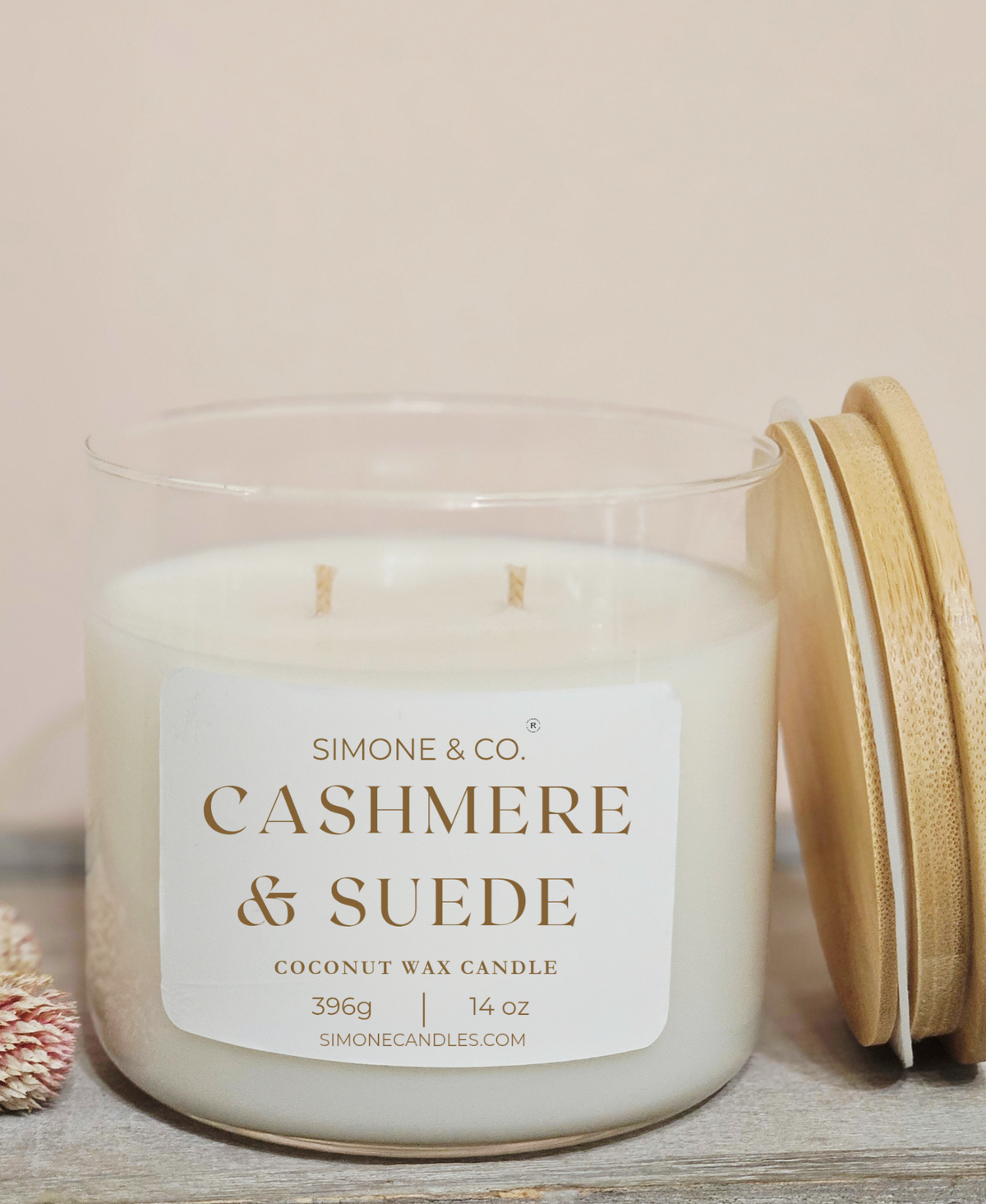 A Simone & Co. 'Cashmere & Suede' coconut wax candle in a clear glass jar with two wicks, shown with its bamboo lid leaning against it on a rustic wooden surface.