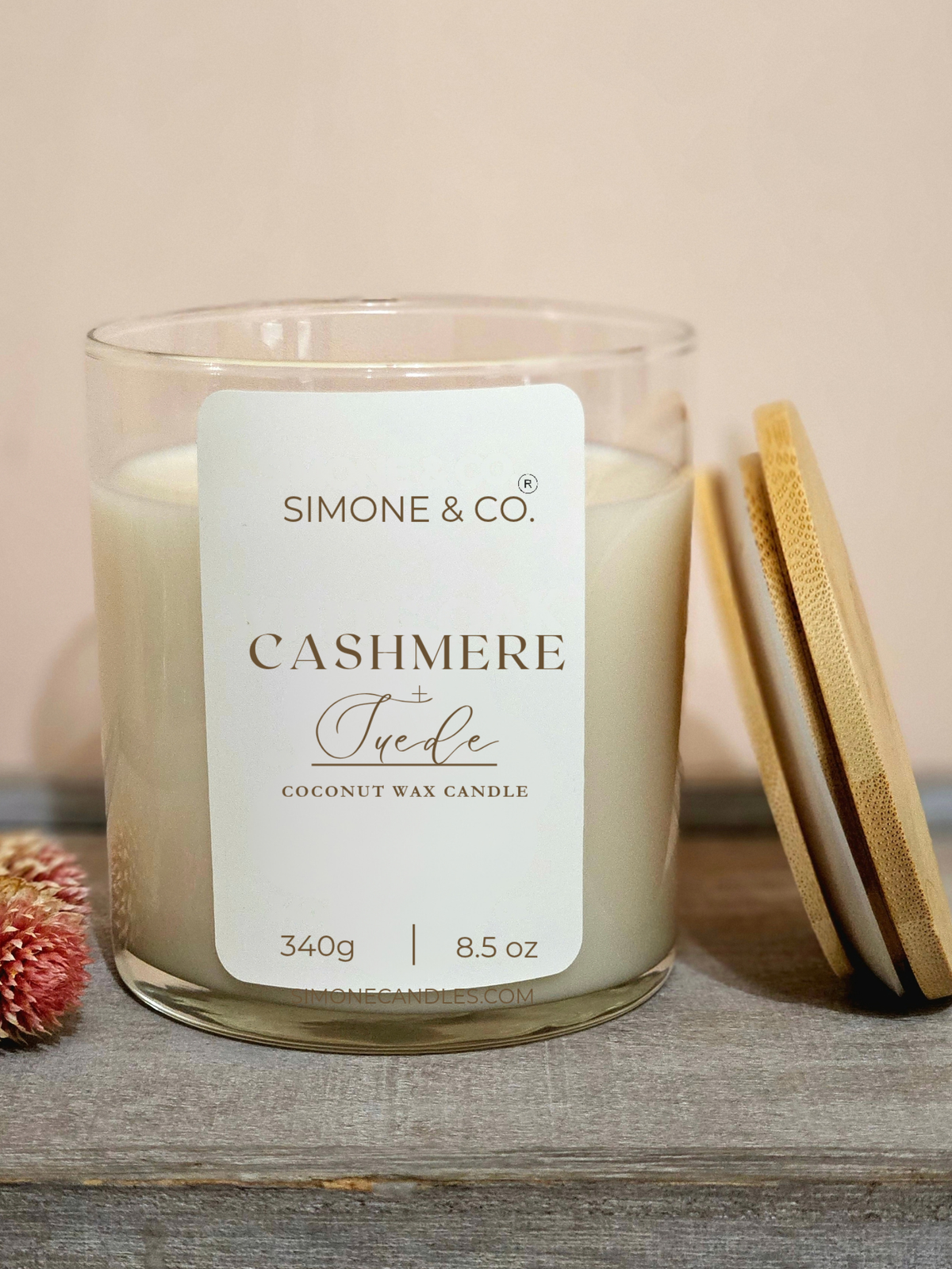 A Simone & Co. Cashmere Jude coconut wax candle in a clear glass jar, shown with its white label and a light wood lid leaning against it on a rustic wooden surface.