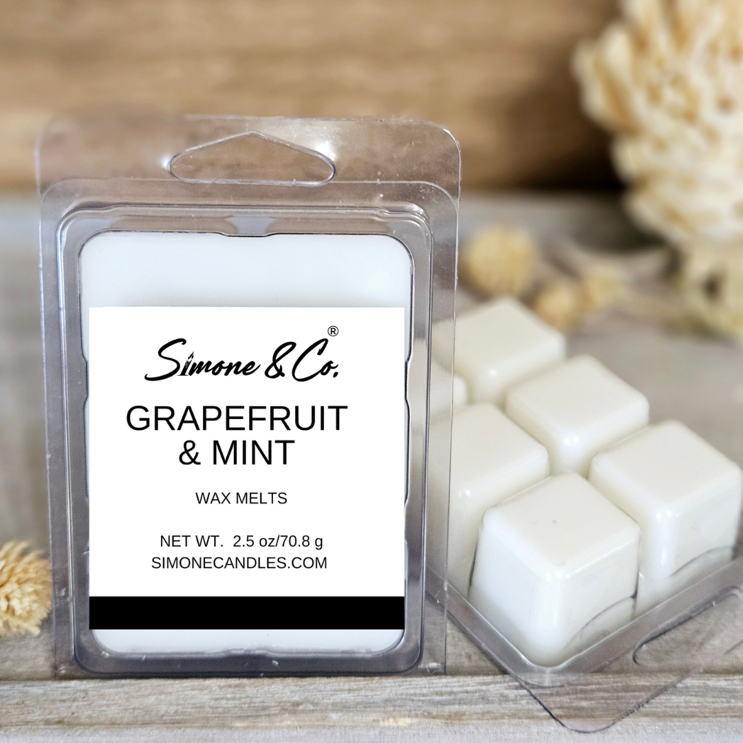 A package of Simone & Co. Grapefruit & Mint wax melts with multiple white wax melt cubes visible, set on a wooden surface with decorative elements in the background.