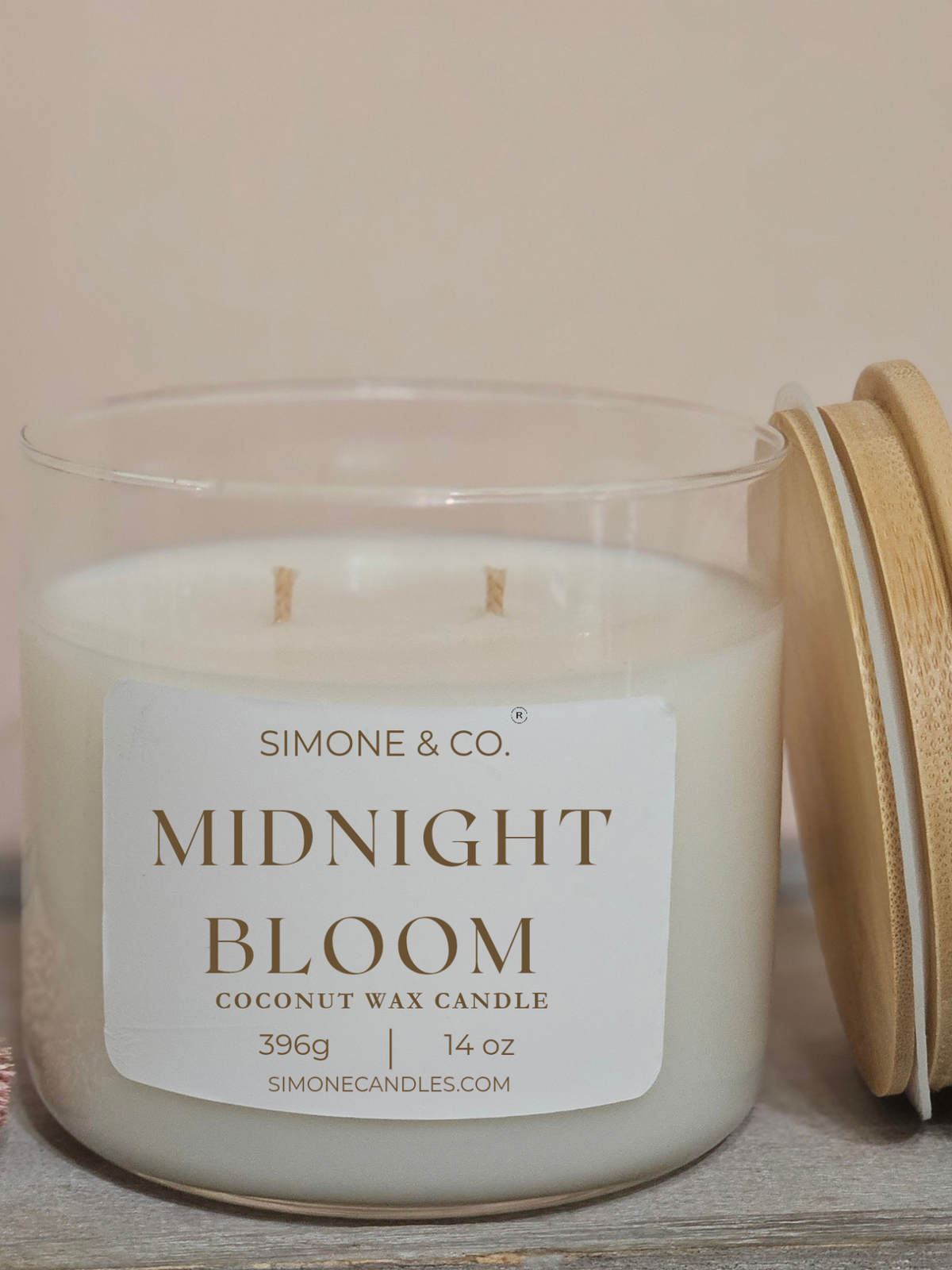 A Simone & Co. 'Midnight Bloom' coconut wax candle in a clear glass jar with a white label and a light wood lid resting beside it on a rustic wooden surface.