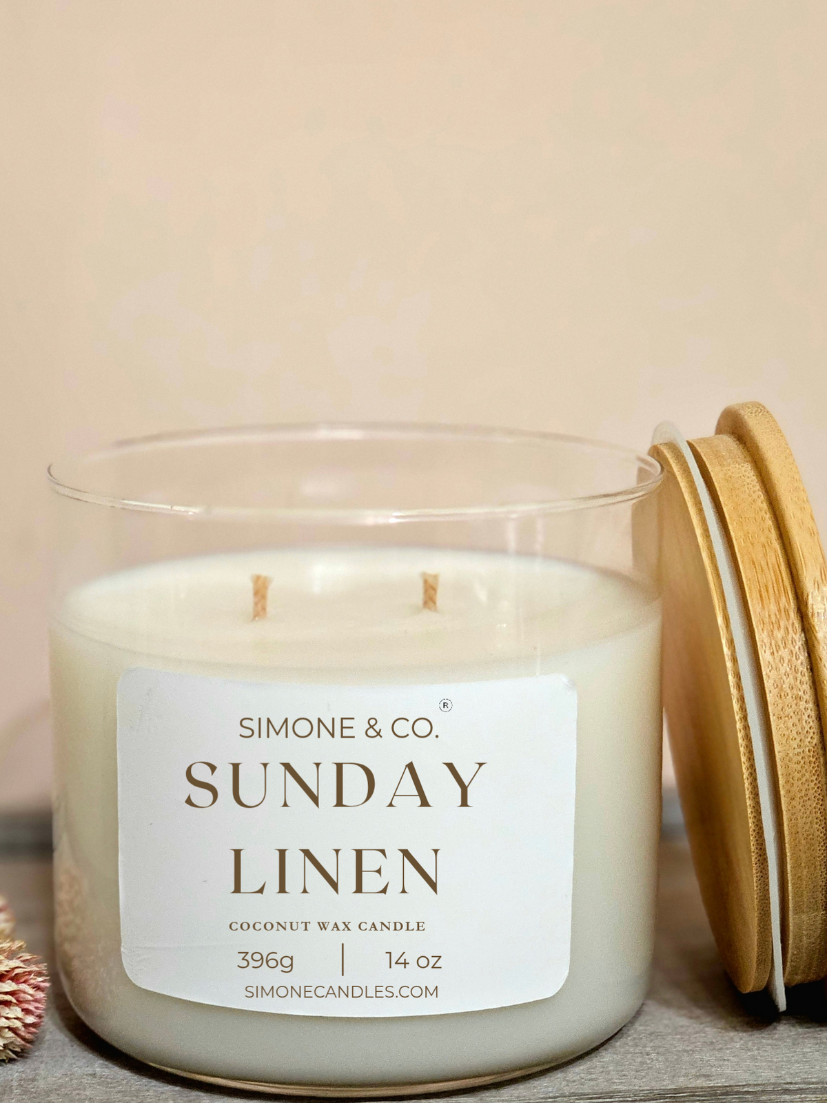 A Simone & Co. 'Sunday Linen' coconut wax candle in a clear glass jar with a white label, shown next to its light wood lid on a neutral beige background.